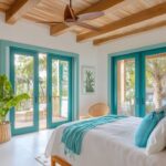 a master bedroom in a tropical-themed house. The bedroom features glass sliding doors with vibrant teal frames that lead out to a deck, providing a seamless connection to the outdoors. The room is designed with a soft, calming color palette that includes crisp white walls and ceilings, with exposed beams at an angle. The ceiling features wooden accents in a light honey tone, matching the overall aesthetic of the house. The bed is dressed in light, airy linens with hints of turquoise and seafoam green, echoing the tropical surroundings. Cozy rattan furniture pieces, such as a reading chair and a small table, add to the inviting atmosphere. Lush green plants are placed strategically around the room to bring in a touch of nature. The flooring is covered in warm, natural stone tile, creating a harmonious feel with the tropical theme. The room is filled with natural light, enhancing the open and airy ambiance. Despite its simplicity, the bedroom is stylish and makes excellent use of space, creating a cozy and inviting atmosphere. --v 6.1 Job ID: e14a5783-5399-4b17-baa3-6997dfc4eceb